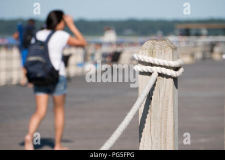 Shallow depth of field looking past railing post overlooking boardwalk along beautiful summer beach. Poeple walk and sit to view scenery and enjoy war Stock Photo