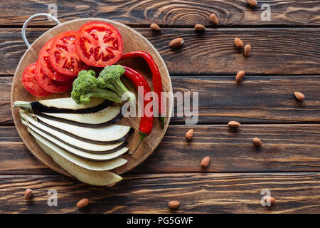 top view of fresh cut eggplant, tomatoes, broccoli and chili peppers on cutting board on wooden tabletop with almonds around Stock Photo