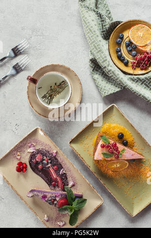 flat lay with sweet carrot cake with berry filling, blueberry cake served with mint leaves and violet petals, cup of herbal tea and cutlery on grey ta Stock Photo