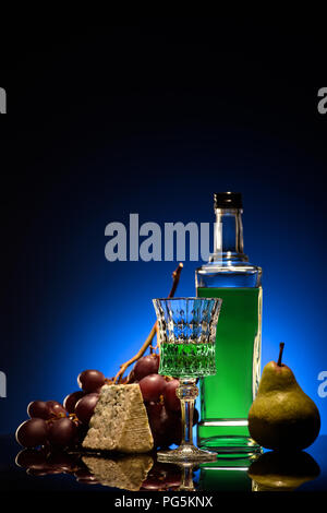 close-up shot of absinthe with ripe pear, grapes and cheese on mirror surface on dark blue background Stock Photo