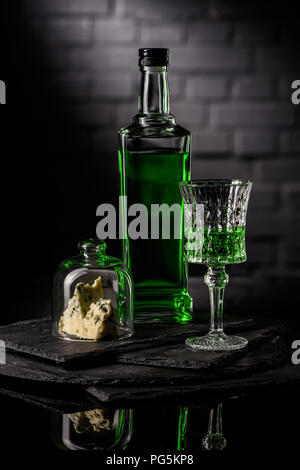 close-up shot of glass and bottle of absinthe with slice of cheese on dark brick wall background Stock Photo