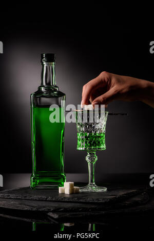 cropped shot of woman putting sugar cube on absinthe spoon on glass on dark background Stock Photo