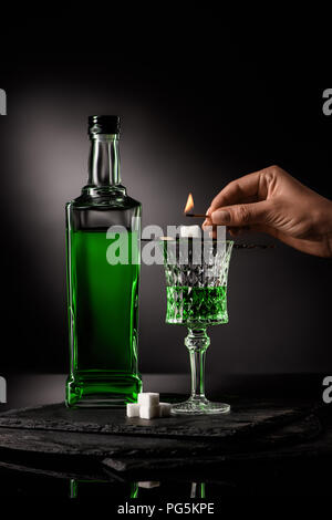 cropped shot of woman holding match over spoon with sugar cube on absinthe glass on dark background Stock Photo