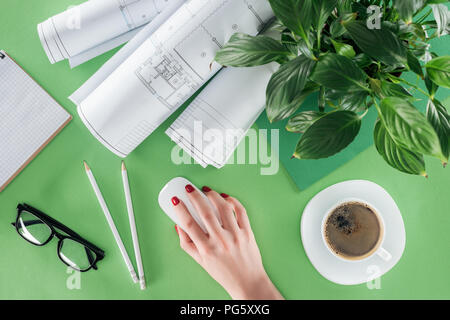 cropped image of female architect using computer mouse at table with blueprints, coffee and plant Stock Photo