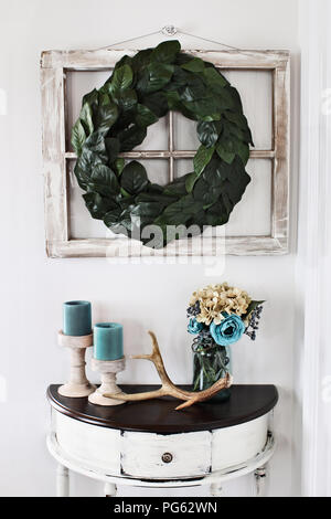 Old farmhouse window decorated with a homemade Magnolia leaf wreath hung on an interior wall over rustic half moon table with candles and flowers deco Stock Photo