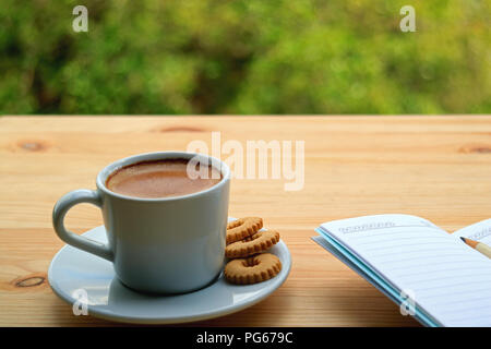 A Cup of Hot Coffee with Cookies and Lined Note Papers on the Wooden Table of Outdoor Seating, Blurred Green Bush in Background Stock Photo