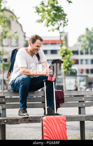 Smiling man with rolling suitcase and takeaway coffee sitting on bench using tablet Stock Photo