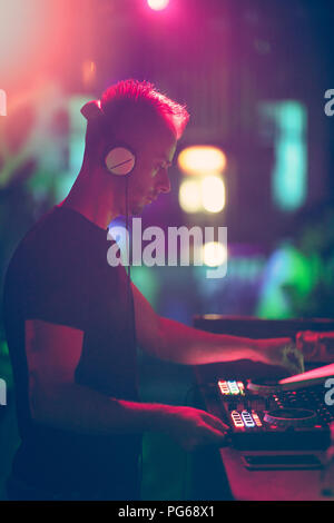 Club, disco DJ playing and mixing music for crowd of happy people. Nightlife, concert lights, flares. Stock Photo