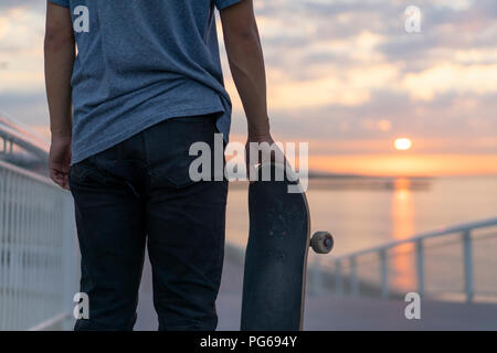 Young man with skateboard at the beach at sunrise, rear view Stock Photo