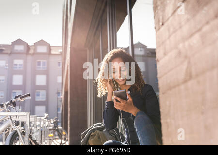 Young woman sitting in front of window in the city, using smartphone Stock Photo