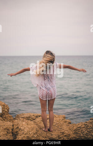 Rear view of young woman standing with outstretched arms on a rock at the coast Stock Photo