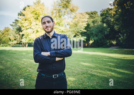 Young businessman standing in park with arms crossed Stock Photo