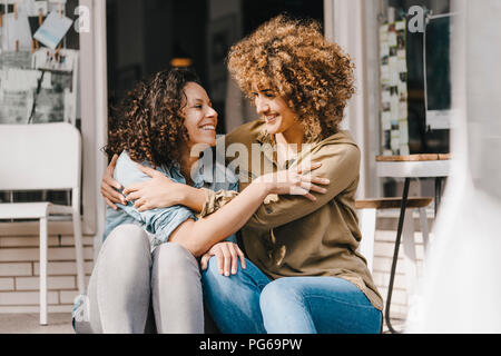 Two laughing friends sitting in front of coworking space, embracing Stock Photo