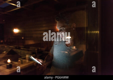 Man leaning at the window in a dark illuminated room using cell phone Stock Photo