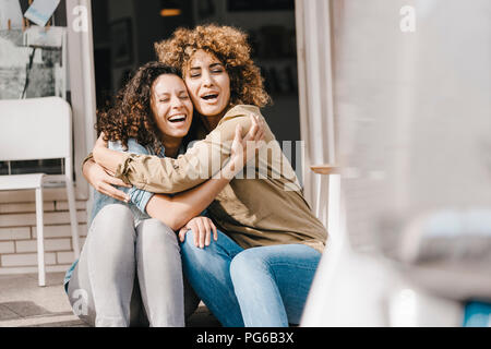 Two laughing friends sitting in front of coworking space, embracing Stock Photo