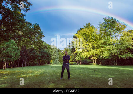 Young businessman standing in park under a rainbow, with arms crossed Stock Photo