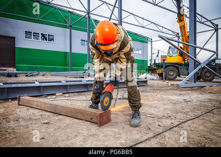 A young  man welder in brown uniform, a construction helmet and welders leathers, grinder metal an angle grinder  at the construction site, orange spa Stock Photo