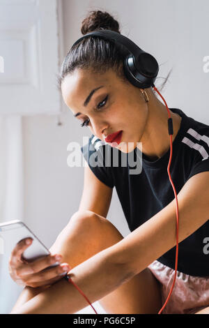 Beautiful young woman sitting down wearing headphones looking at cell phone Stock Photo