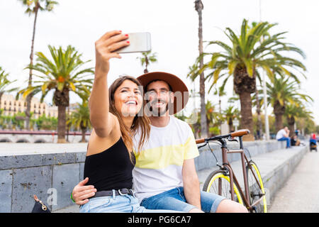 Spain, Barcelona, happy couple sitting on bench taking a selfie Stock Photo