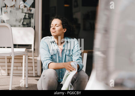 Young woman sitting in front of coffee shop with eyes closed, enjoying the sun Stock Photo