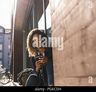 Young woman sitting in front of window in the city, using smartphone Stock Photo