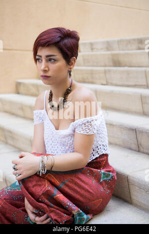 Beautiful young woman sat on the steps outdoors looking away Stock Photo