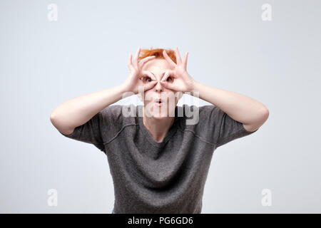 Cheerful mature woman with red hair holding fingers near eyes like glasses Stock Photo