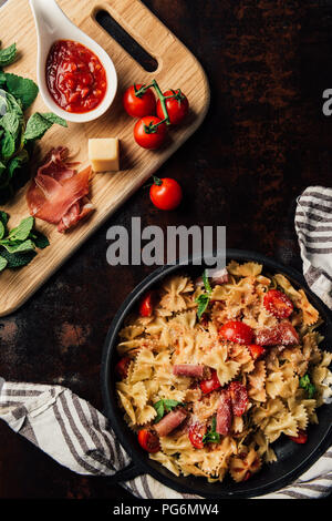 top view of pasta with jamon, pine nuts, sauce, cherry tomatoes, mint leaves covered by grated parmesan in pan surrounded by kitchen towel and cutting Stock Photo