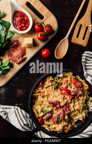 elevated view of pasta with jamon, pine nuts, sauce, cherry tomatoes, mint leaves covered by grated parmesan in pan surrounded by kitchen towel and cu Stock Photo