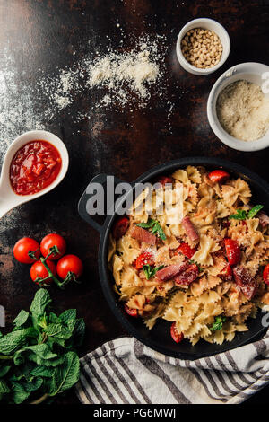view from above of pasta with jamon, pine nuts, sauce, cherry tomatoes, mint leaves covered by grated parmesan in pan surrounded by ingredients and ki Stock Photo