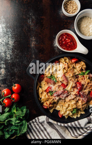 elevated view of pasta with jamon, pine nuts, sauce, cherry tomatoes, mint leaves covered by grated parmesan in pan surrounded by ingredients and kitc Stock Photo