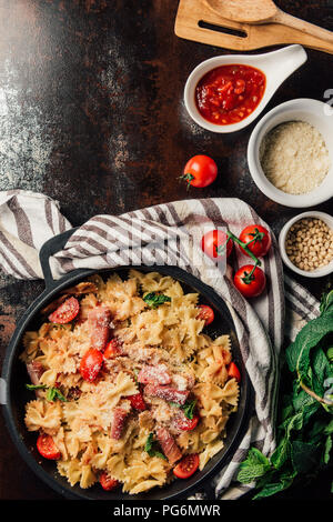 top view of pasta with jamon, pine nuts, sauce, cherry tomatoes, mint leaves covered by grated parmesan in pan surrounded by ingredients and kitchen t Stock Photo
