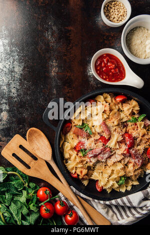 elevated view of pasta with jamon, pine nuts, sauce, cherry tomatoes, mint leaves covered by grated parmesan in pan surrounded by ingredients and kitc Stock Photo