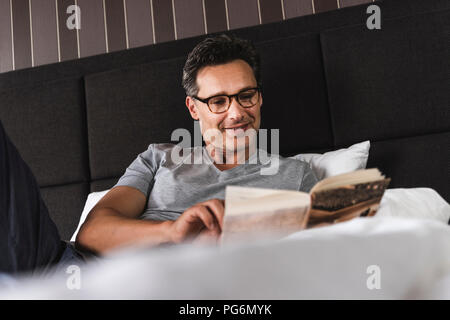 Smiling man lying in bed at home reading a book Stock Photo