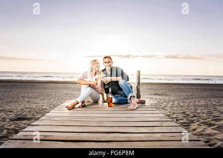 Happy couple with wine glasses sitting on boardwalk on the beach at sunset Stock Photo