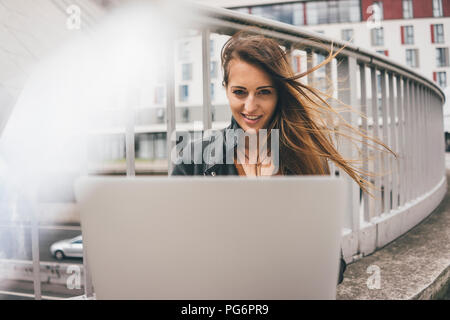 Portrait of smiling young woman with windswept hair using laptop on motorway bridge Stock Photo
