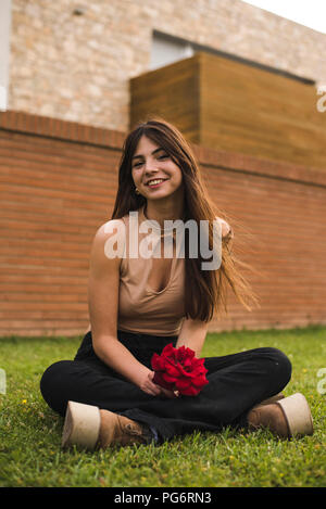 Portrait of smiling young woman with red rose sitting on lawn Stock Photo