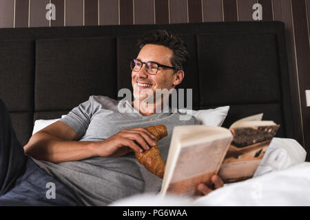 Happy man lying in bed at home with croissant and book Stock Photo