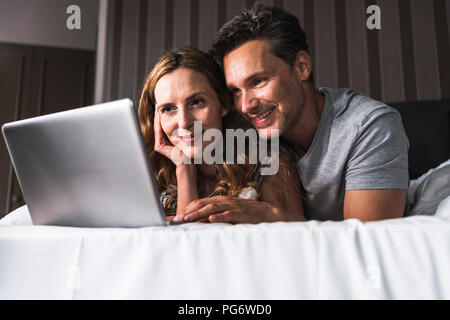 Smiling couple lying on bed at home looking at laptop Stock Photo