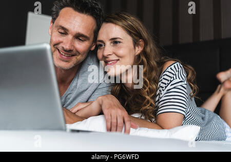 Happy couple lying on bed at home looking at laptop Stock Photo