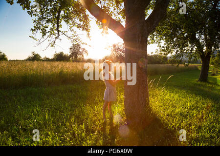 Young girl standing at tree against evening sun in summer Stock Photo