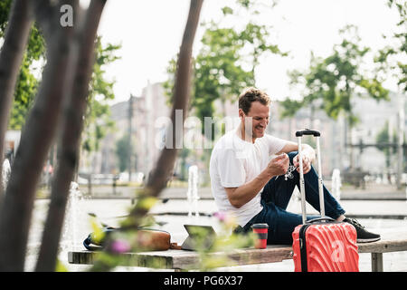 Smiling man with rolling suitcase sitting on bench Stock Photo