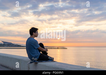 Young Chinese man with skateboard sitting on wall at the beach Stock Photo