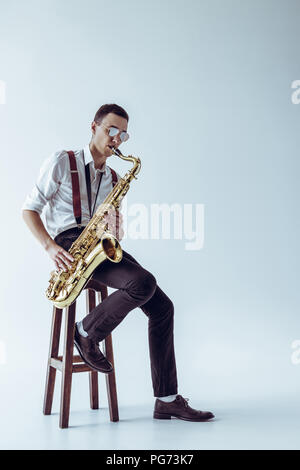 handsome young performer sitting on stool and playing saxophone on grey Stock Photo