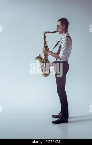 side view of stylish young professional musician playing saxophone on grey Stock Photo