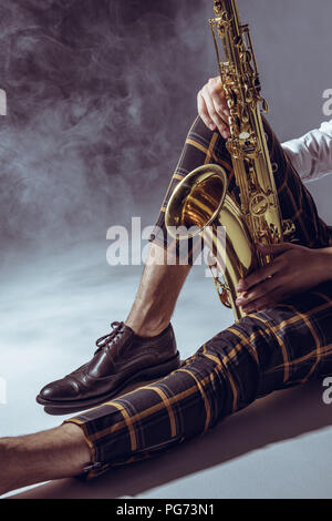 partial view of stylish young musician sitting with saxophone in smoke on grey Stock Photo