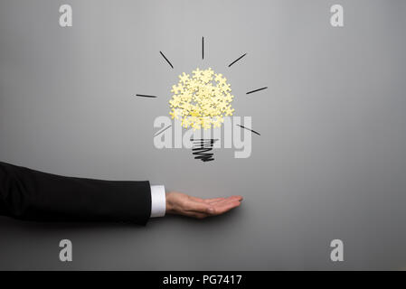 Conceptual bulb drawing with yellow scattered puzzle pieces over open businessman hand on gray background. Stock Photo