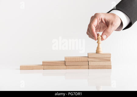 Businessman placing queen chess piece on a stepped stack of wooden blocks on the highest level conceptual of success. Stock Photo