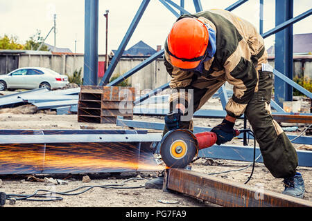 A young  man welder in brown uniform, a construction helmet and welders leathers, grinder metal an angle grinder  at the construction site, orange spa Stock Photo