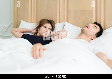 Annoyed Asian wife blocking her ears from noise of husband snoring in bedroom at home. Young couple have problem with man's snoring. Stock Photo